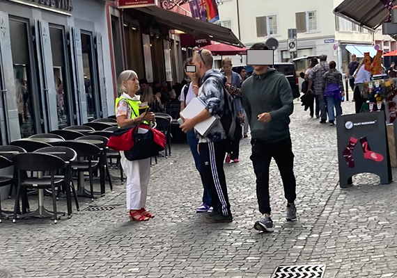 Tag des Weissen Stockes Aktion in Zürich - Blindversuche. Eine betroffene Person mit gelber Weste spricht zwei Passanten an und verteilt ihnen Infomaterial.