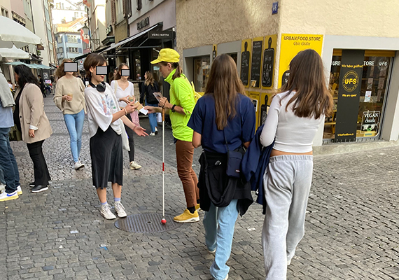 Tag des Weissen Stockes Aktion in Zürich - Blindversuche. Eine betroffene Person mit gelber Weste und gelbem Blindenbund-Käppli und Weissem Stock spricht eine Passantin an und verteilt ihr Infomaterial.