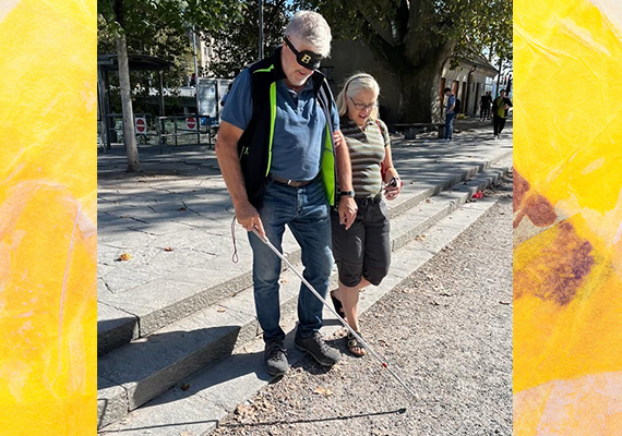 Tag des Weissen Stockes Aktion in Zürich - Blindversuche. Eine männliche Person mit Dunkelbrille geht mit dem Weissen Stock und geht eine kleine Treppe hinab. Neben ihm ist eine Begleiterin, die ihn am Arm führt.