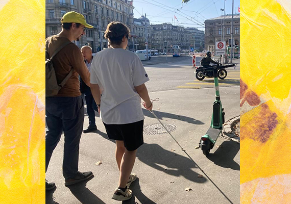 Tag des Weissen Stockes Aktion in Zürich - Blindversuche. Eine Person in weissem T-Shirt geht mit dem Weissen Stock und kommt einem E-Trotti nahe. Neben ihr ist ein Begleiter mit gelbem Blindenbund-Käppli